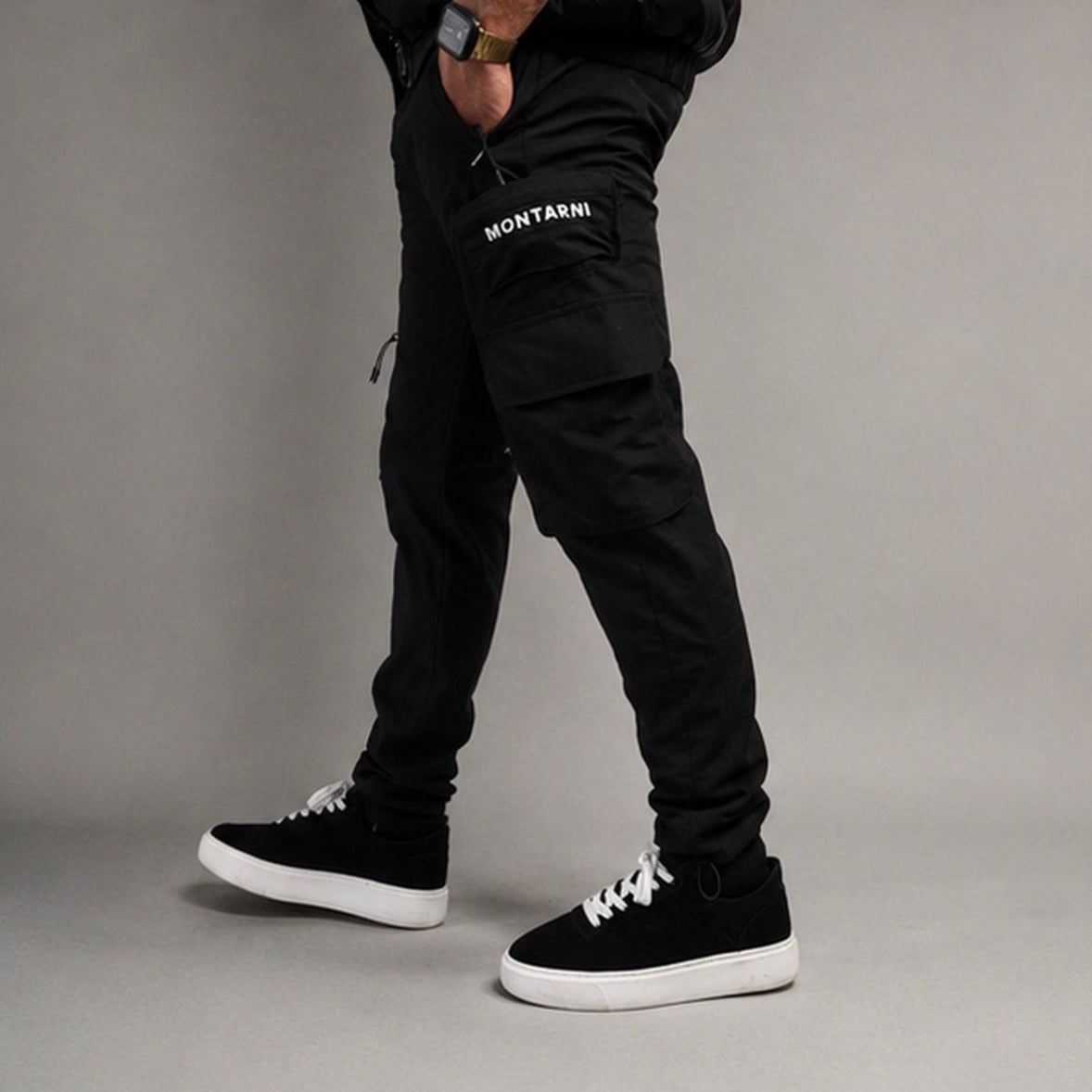 A person wearing black Montarni cargo jogger pants and black high-top sneakers with white laces and white soles, standing against a plain gray studio background.